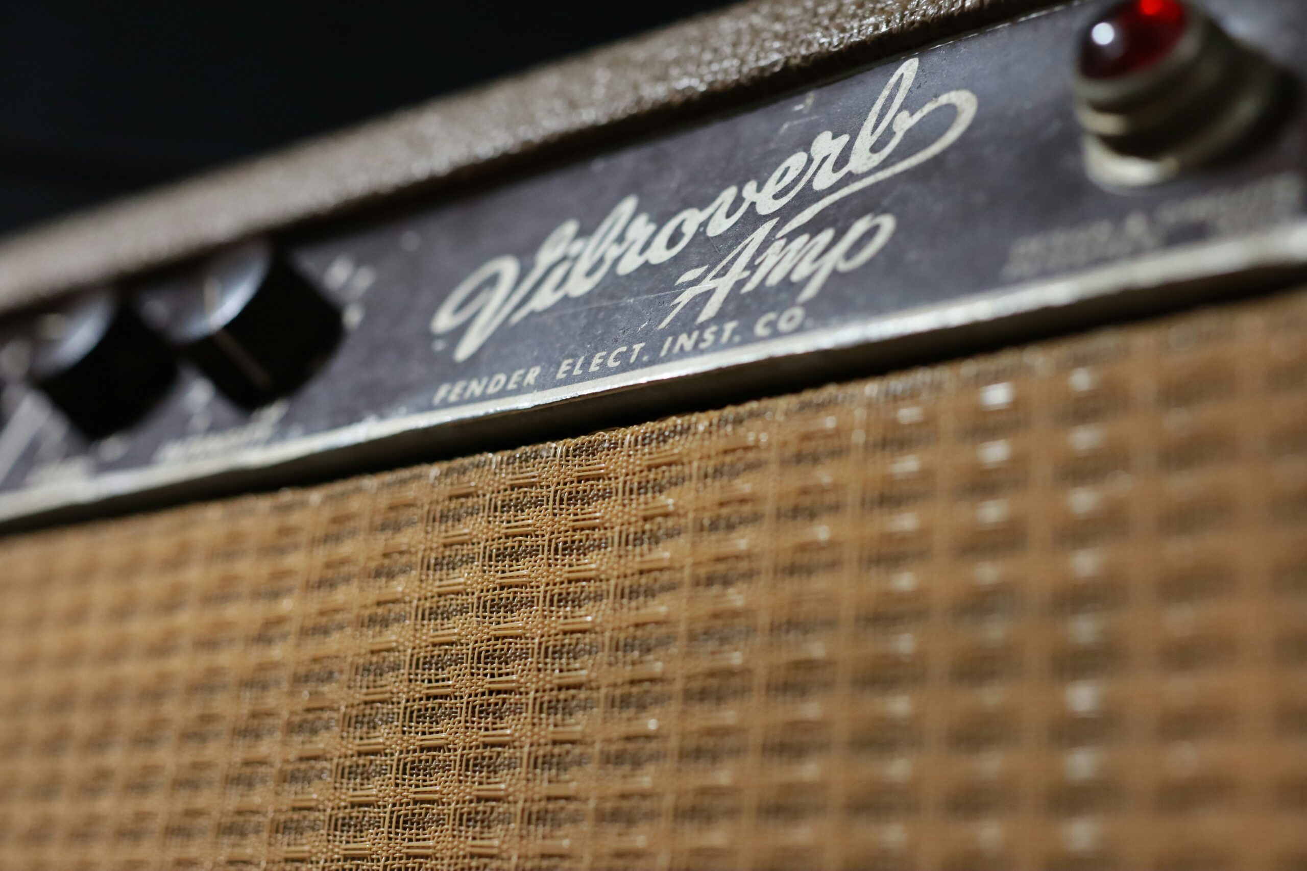 Close-up of a vintage Fender Vibroverb guitar amplifier, showing the textured brown speaker grille, black control knobs, and the worn metallic nameplate with cursive branding.